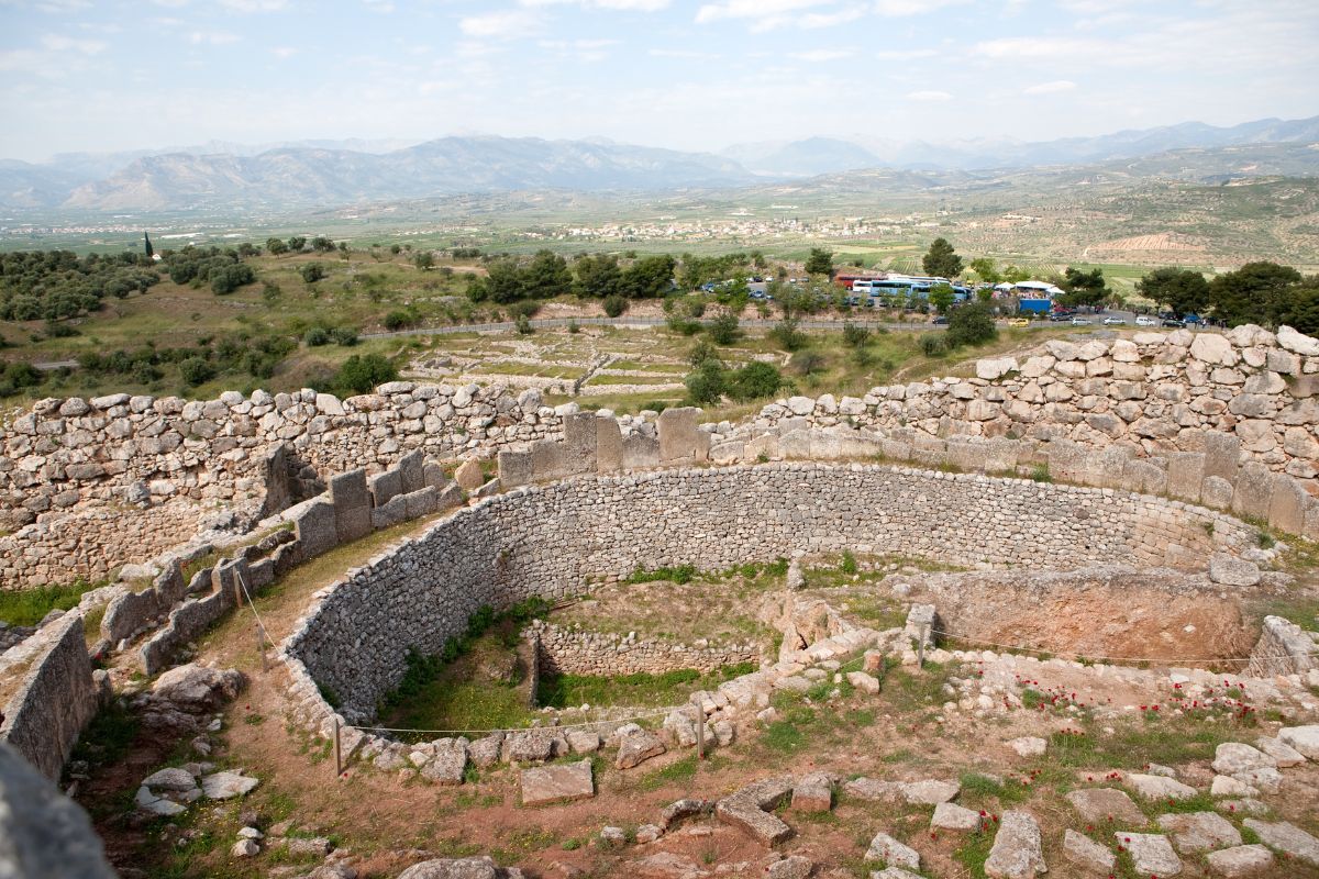 The image shows an aerial view of the ancient Mycenaean archaeological site, featuring circular stone structures and surrounding landscape with distant mountains.