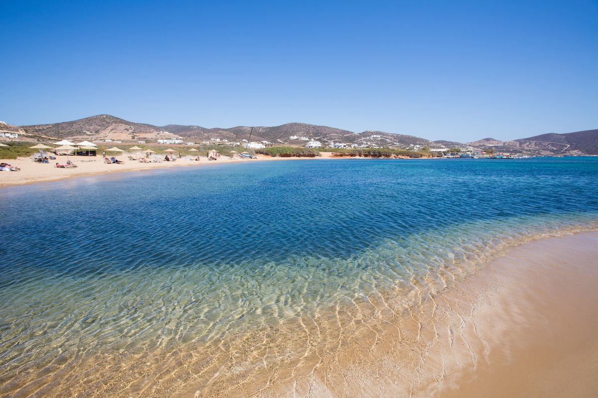 The image shows a serene beach on Antiparos, featuring clear, shallow waters gently lapping against a sandy shoreline. The beach is backed by rolling hills and scattered sunbathers, offering a peaceful and picturesque setting typical of the Greek island's natural beauty.