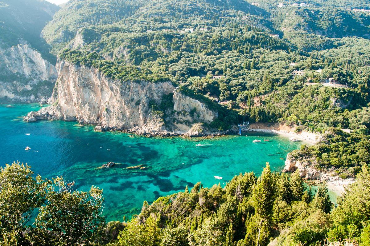 A scenic view of Corfu showcasing lush green hills and dramatic cliffs surrounding a turquoise bay with clear waters and small boats.