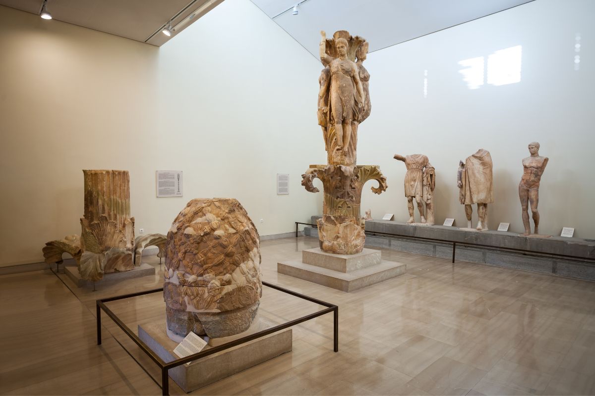 Interior of the Delphi Archaeological Museum, showcasing various ancient statues and artifacts, including a large column capital and a stone sculpture of the omphalos.