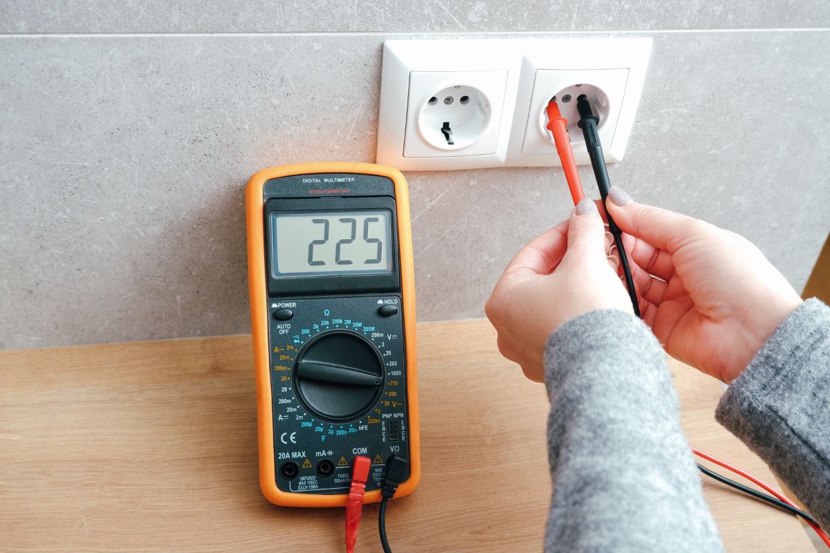 A person using a digital multimeter to measure the voltage of an electrical outlet, displaying a reading of 225 volts.