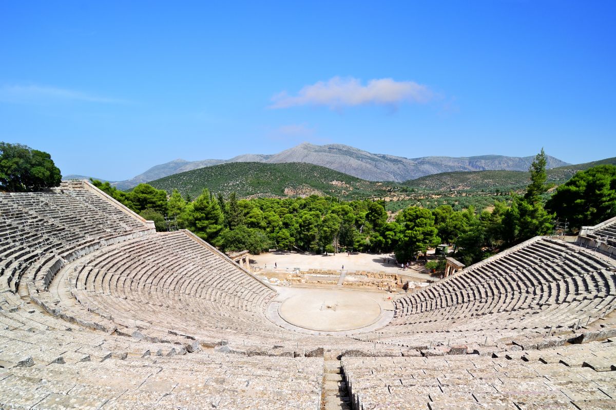 The ancient theater of Epidaurus, surrounded by lush greenery and mountainous landscape, features stone seating arranged in a semi-circular layout, showcasing its architectural brilliance and historical significance.