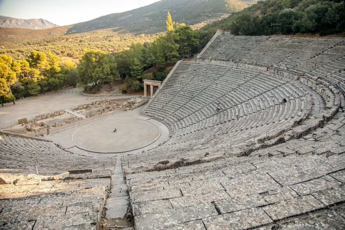 A large ancient amphitheater, Epidaurus in Greece, partially surrounded by lush green hills, showcasing the grand stone seating and central performance area.