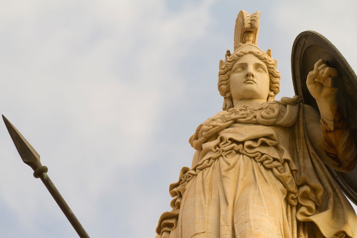 Statue of the goddess Athena holding a spear and shield, viewed from below against a cloudy sky.