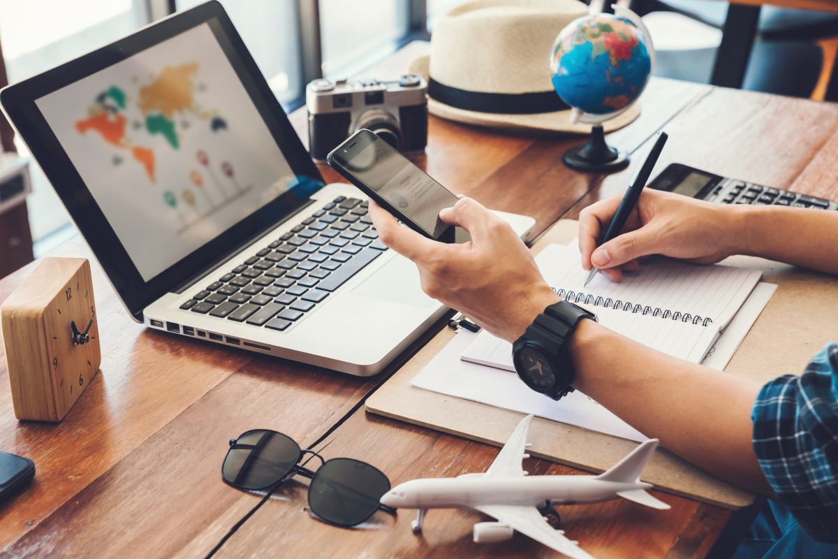 A workspace setup with a laptop, smartphone, notebook, miniature globe, and travel-related items, suggesting travel planning or itinerary preparation.