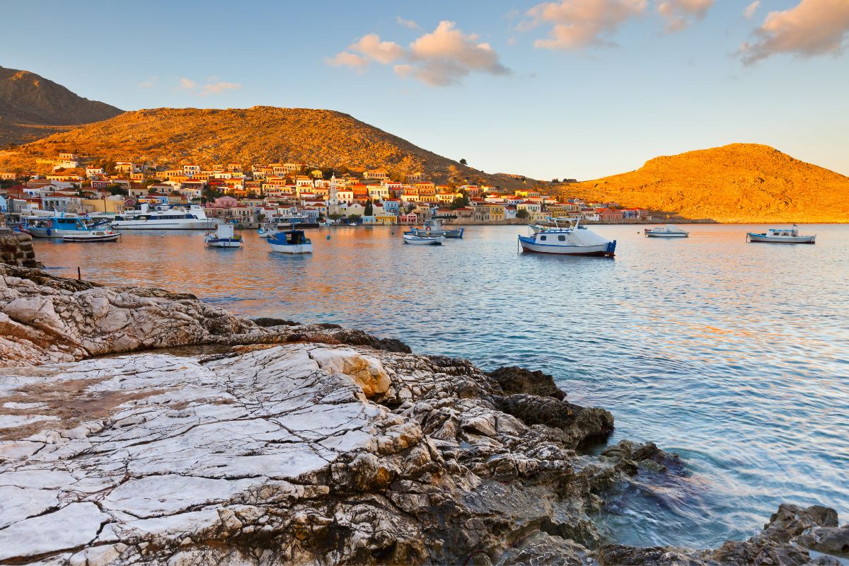 A picturesque view of Halki Village in Naxos, with colorful houses nestled against sunlit hills, small boats floating in the calm waters of the harbor, and rocky shores in the foreground during a warm, golden sunset.