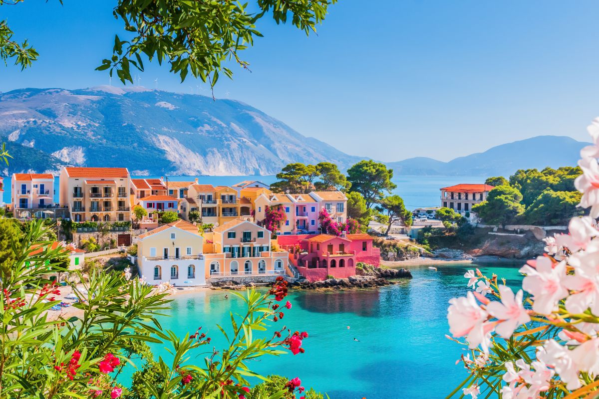 A vibrant seaside village in Kefalonia, featuring colorful houses, turquoise waters, and lush greenery under a clear blue sky.