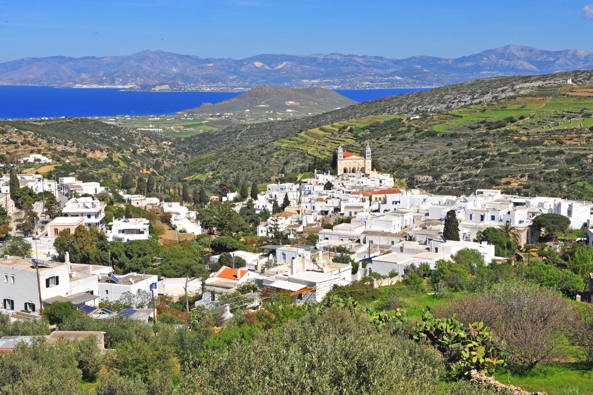The image shows Lefkes village in Paros, nestled among green hills with traditional white-washed buildings. A prominent church stands in the center, and the village overlooks the distant blue waters of the Aegean Sea, highlighting the serene and picturesque charm of the area.