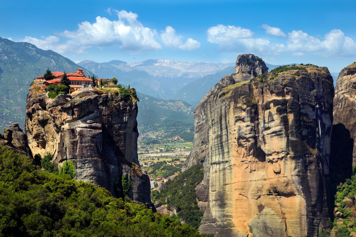 A panoramic view of Meteora, featuring a monastery perched atop a towering rock formation, surrounded by lush green landscapes and distant mountains under a bright blue sky.