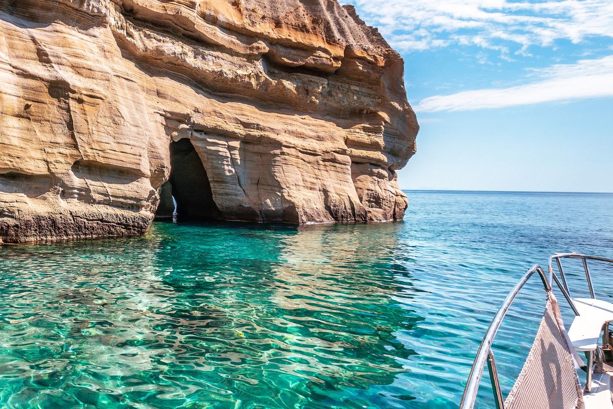 The image shows a rocky cliff formation with a sea cave along the coast of Milos, Greece. The clear turquoise water shimmers under the sunlight, and part of a boat is visible in the foreground, suggesting an exploration of the natural beauty of the island. The rock's layers and textures create a striking contrast against the calm sea.