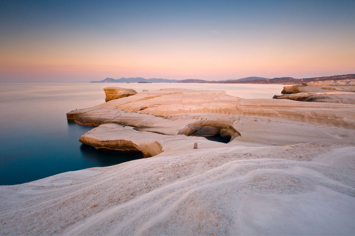 A serene coastal view of Milos Island, showcasing smooth white rock formations meeting calm, clear waters at sunset.