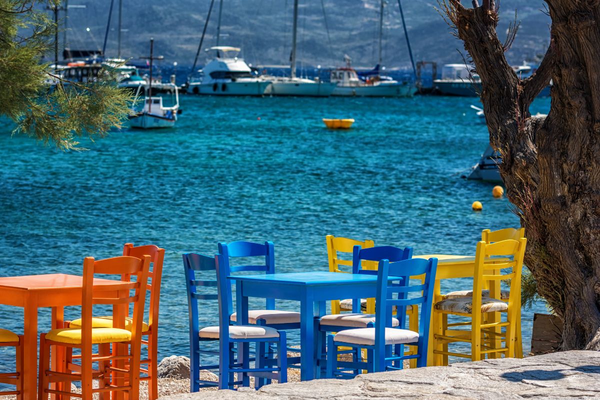 The image shows a vibrant outdoor dining area on Milos Island. The scene is filled with brightly colored tables and chairs, situated by the water's edge with boats visible in the background. The clear, turquoise water contrasts beautifully with the vibrant furniture, creating a picturesque and inviting setting typical of a Greek island.