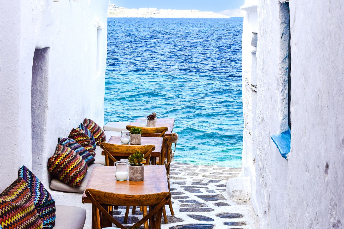 A cozy outdoor cafÃ© in Mykonos, featuring colorful cushions and wooden tables, with a stunning view of the blue Aegean Sea.