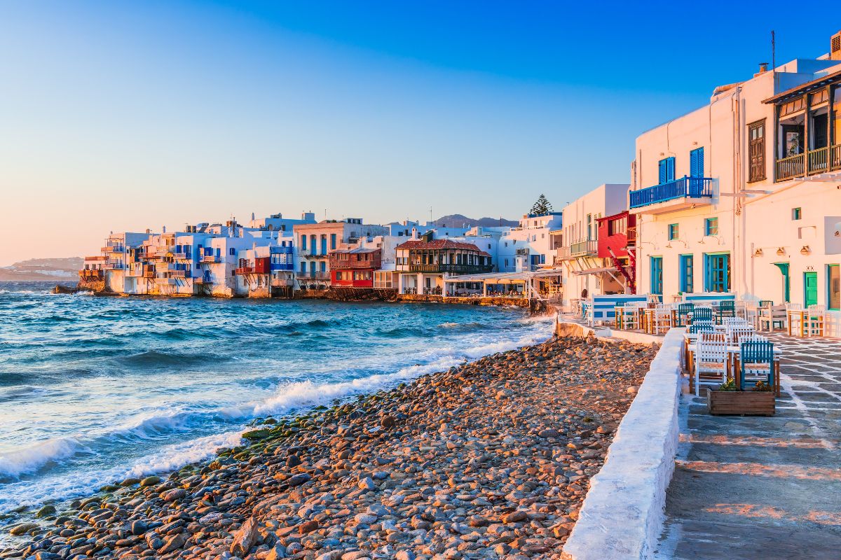 The iconic Little Venice in Mykonos, featuring colorful waterfront houses with balconies overhanging the sea, set against a clear blue sky and waves gently lapping the shore.