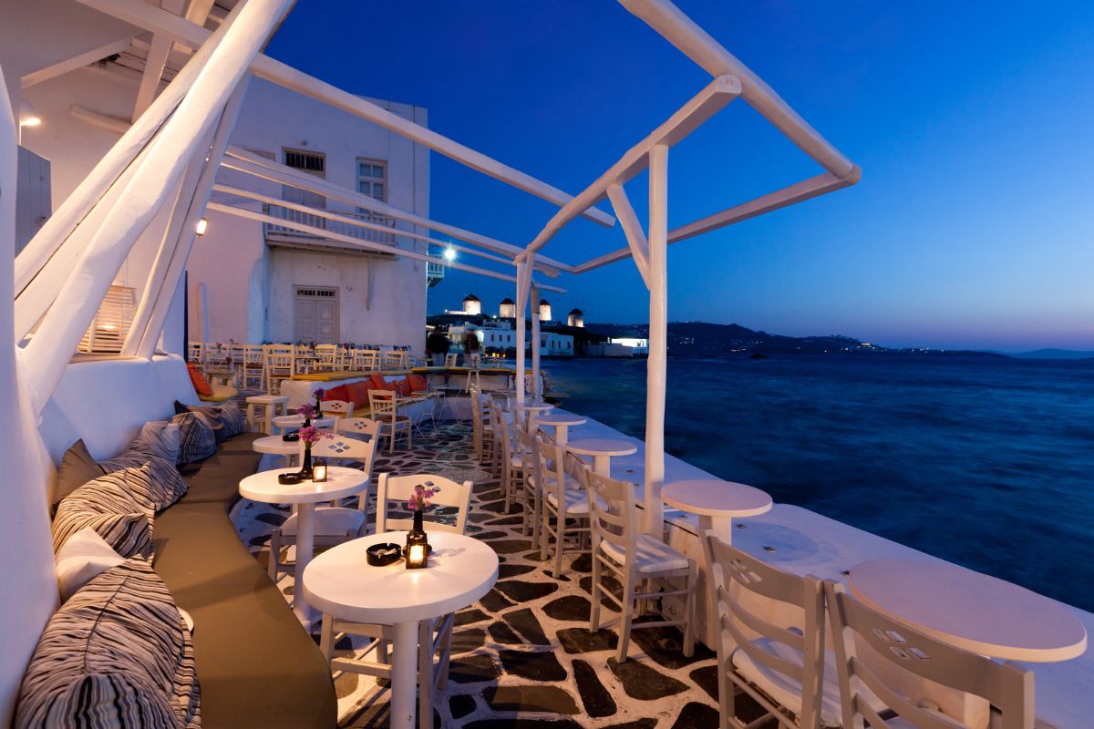 An outdoor cafÃ© in Mykonos at dusk, with cozy seating and a view of the sea under a twilight sky_Mykonos Greece Vacation