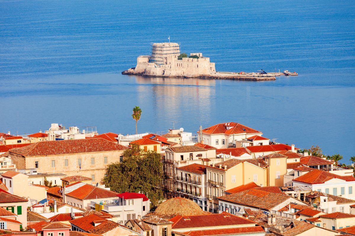 A view of Nafplio featuring charming buildings with red-tiled roofs and a distant fortress sitting on the calm waters, against a backdrop of a serene blue sea.