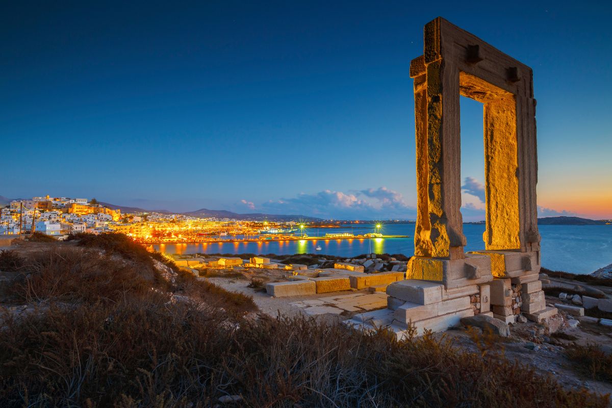 A view of Naxos Island at dusk, featuring the illuminated ancient Portara (temple entrance) in the foreground and the town with its lights reflecting on the water in the background.