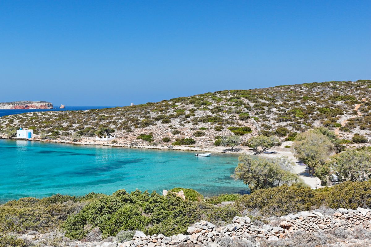 The image shows a tranquil beach on Paros, with clear turquoise waters gently meeting a rocky shoreline. A small white and blue building sits near the water, surrounded by low-lying shrubs and sparse trees, creating a peaceful and secluded coastal scene.