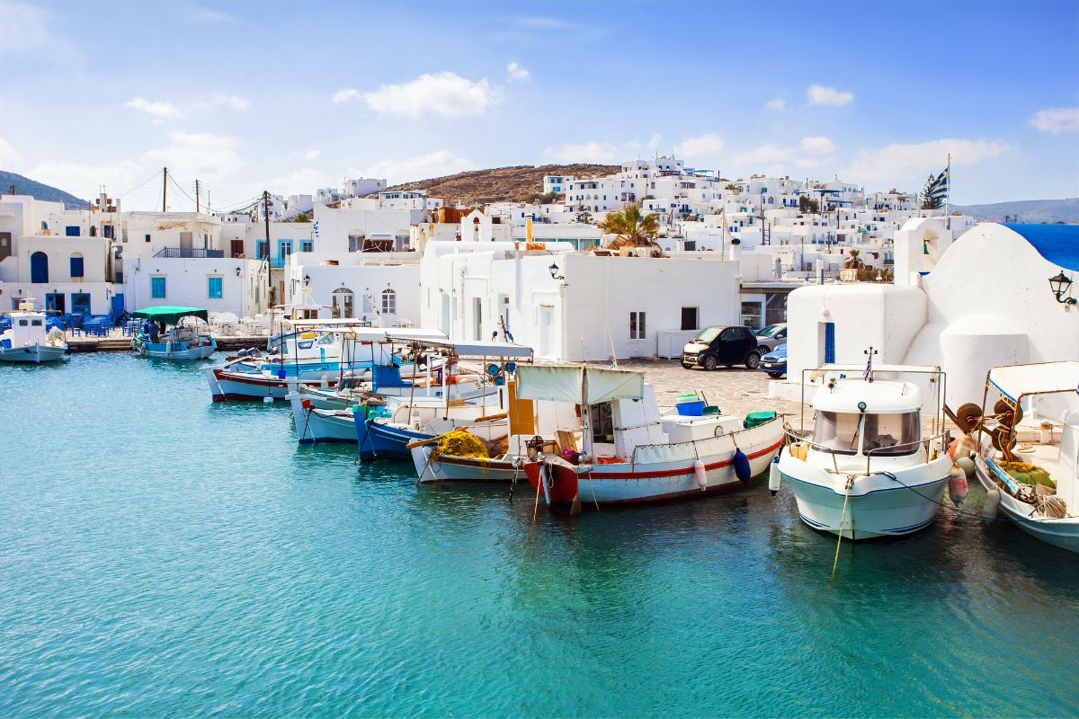 The image shows a charming harbor in Paros, featuring traditional white-washed buildings and colorful fishing boats docked along the clear blue waters. The scene captures the picturesque and serene ambiance of this Greek island.