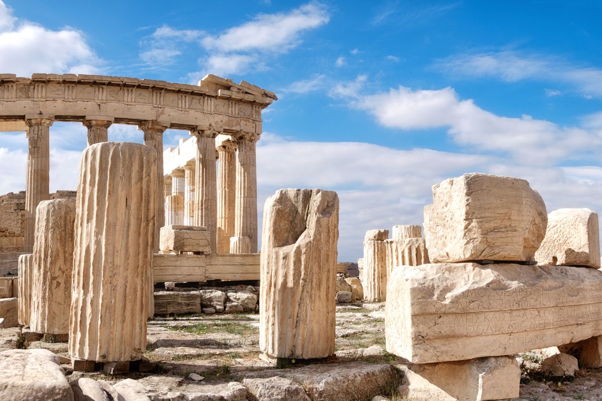 Ruins of the Parthenon in Athens, featuring ancient columns and stone blocks under a bright blue sky with scattered clouds.