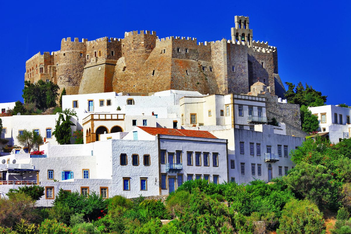 A view of Patmos Island featuring the imposing medieval castle with whitewashed buildings and lush greenery in the foreground under a clear blue sky.