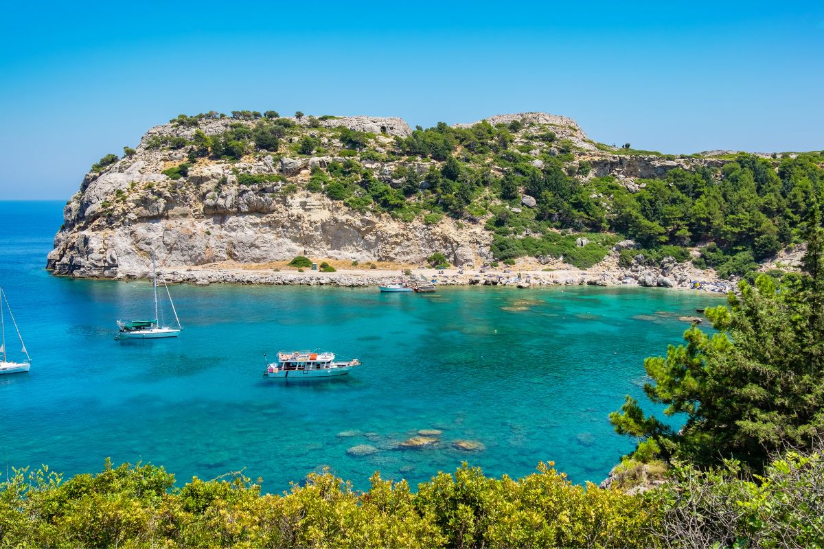 A picturesque view of Rhodes Island featuring clear turquoise waters, a rocky coastline, and lush greenery. Sailboats float peacefully in the calm sea, inviting a sense of serenity and adventure.