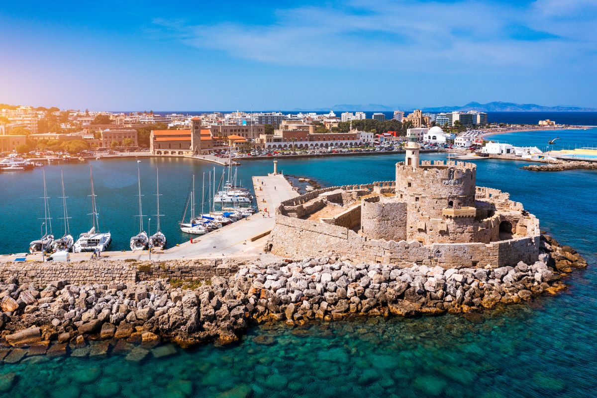 A coastal view of Rhodes, showcasing a historic stone fort surrounded by turquoise waters and a marina with sailboats, under a bright blue sky.