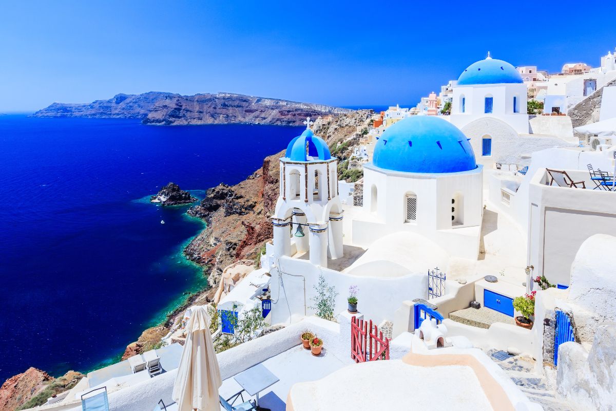 View of Santorini featuring iconic white buildings with blue domes perched on cliffs overlooking the deep blue Aegean Sea, with a stunning coastline in the background.