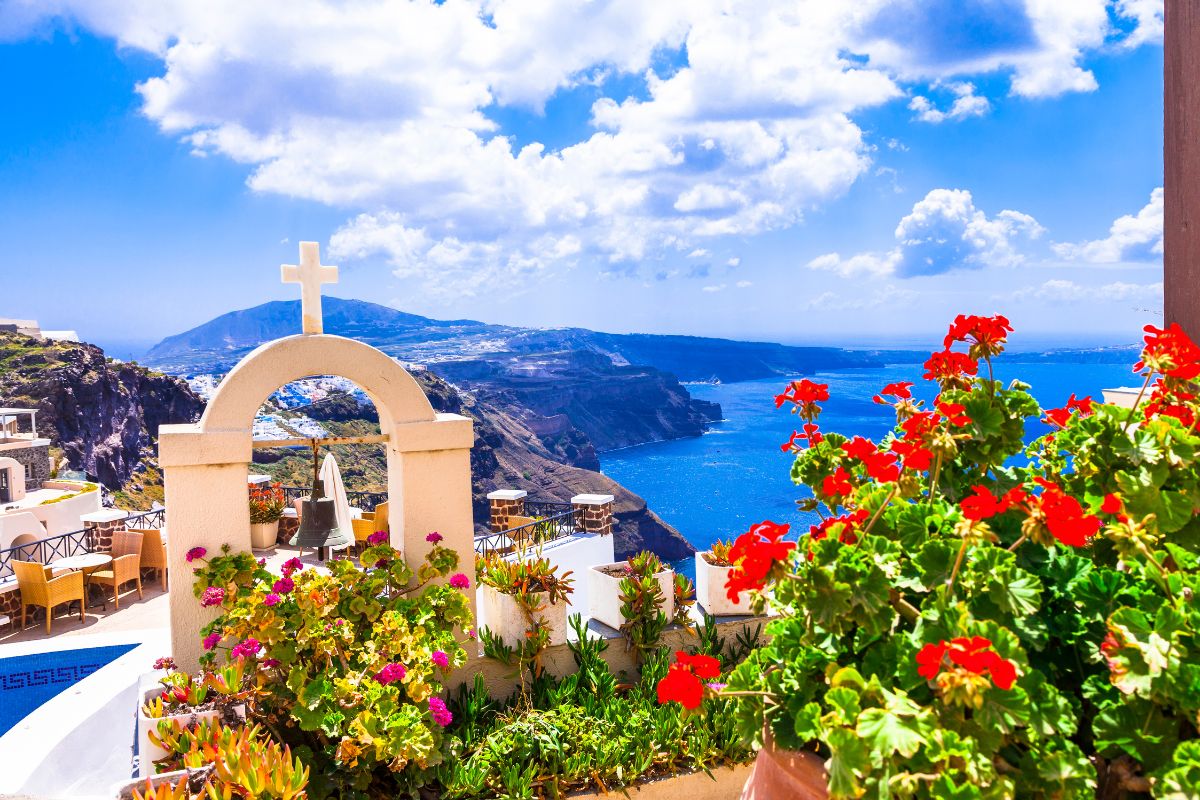 A vibrant scene of blooming flowers in Santorini during spring, with a white archway topped by a cross overlooking the deep blue sea and rocky cliffs under a bright, clear sky.