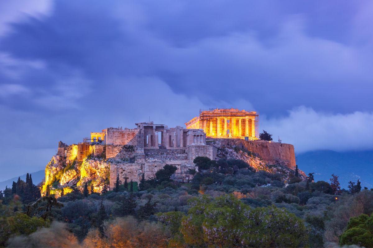The Acropolis Hill in Athens, illuminated at dusk with the Parthenon and surrounding ancient structures glowing against a dramatic sky.
