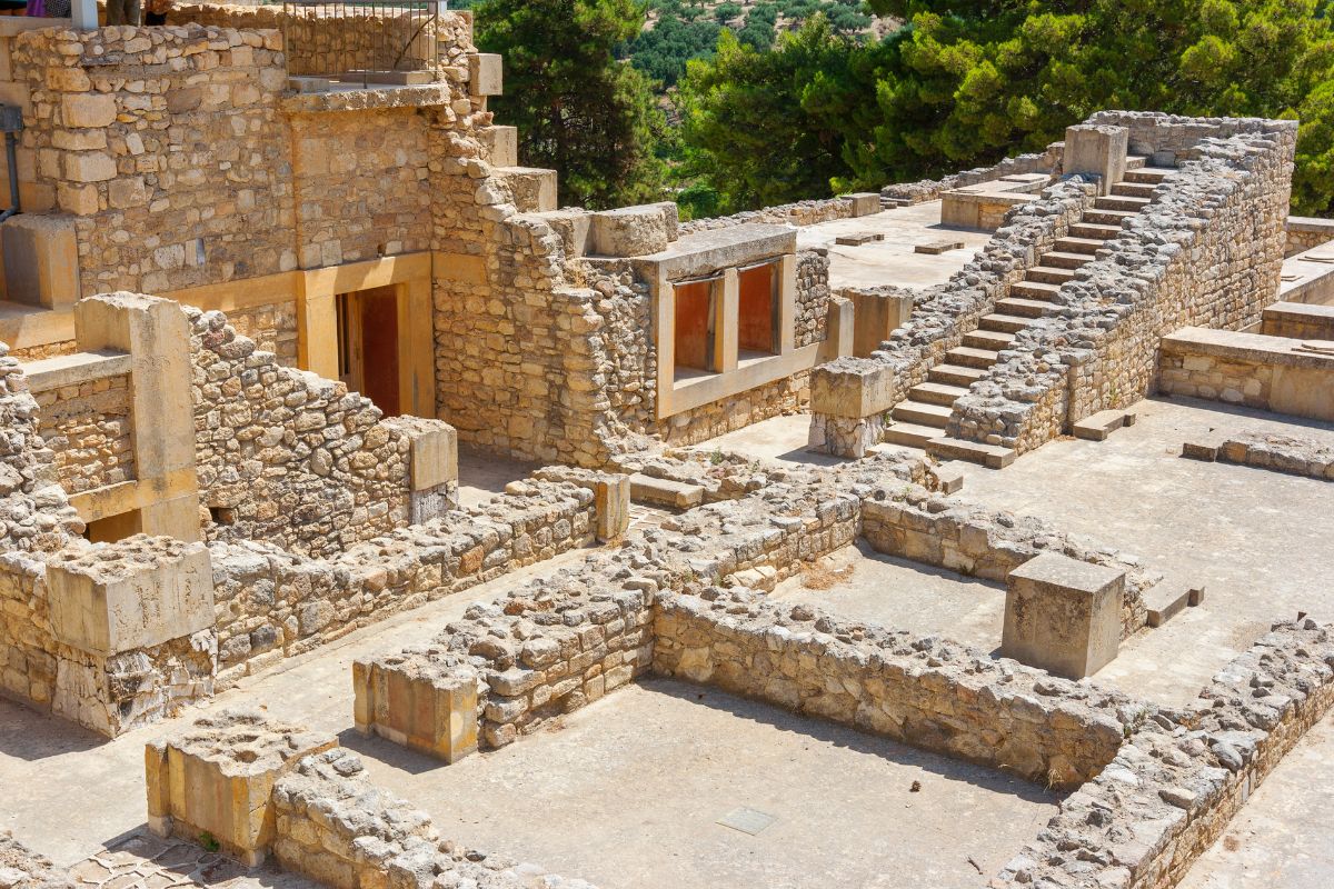 The image shows the archaeological site of Knossos Palace, featuring stone walls, staircases, and remnants of ancient structures amidst a lush green landscape.