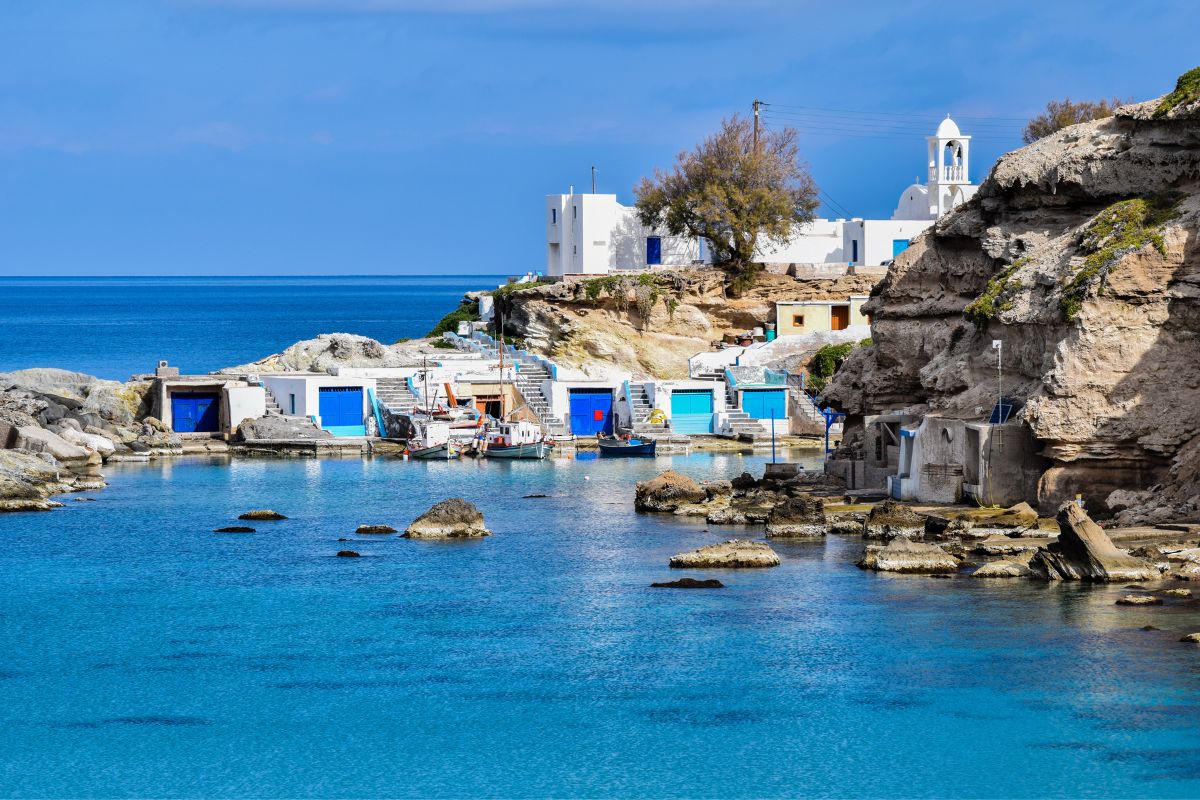 The image shows a peaceful coastal scene in Milos, Greece, featuring traditional whitewashed houses with blue doors and small fishing boats docked in a calm, rocky bay. The clear blue water contrasts with the rugged cliffs, creating a serene and picturesque atmosphere. A small church with a bell tower is visible in the background.