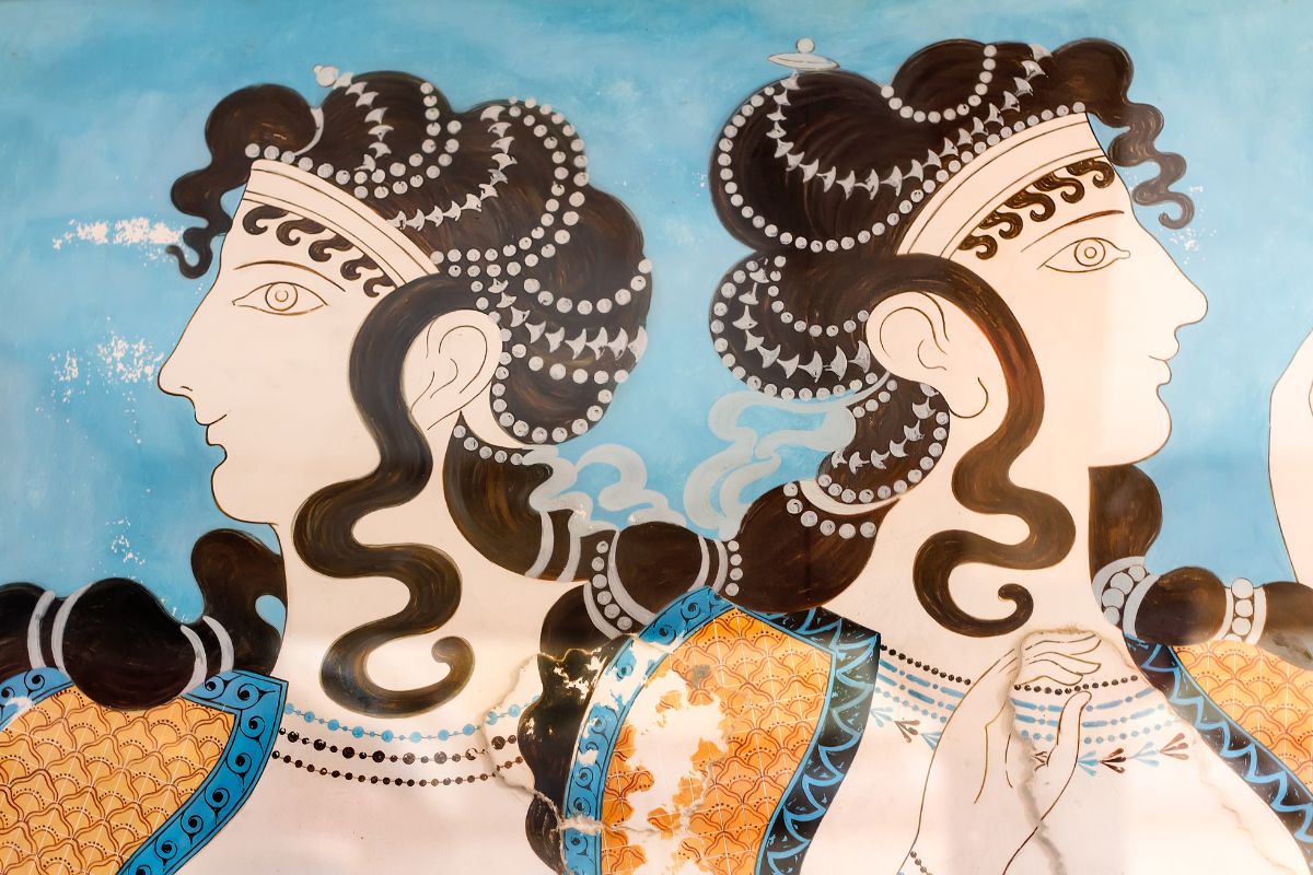 The image depicts a Minoan fresco at Knossos Palace, featuring two women with elaborate hairstyles and vibrant clothing against a blue background.