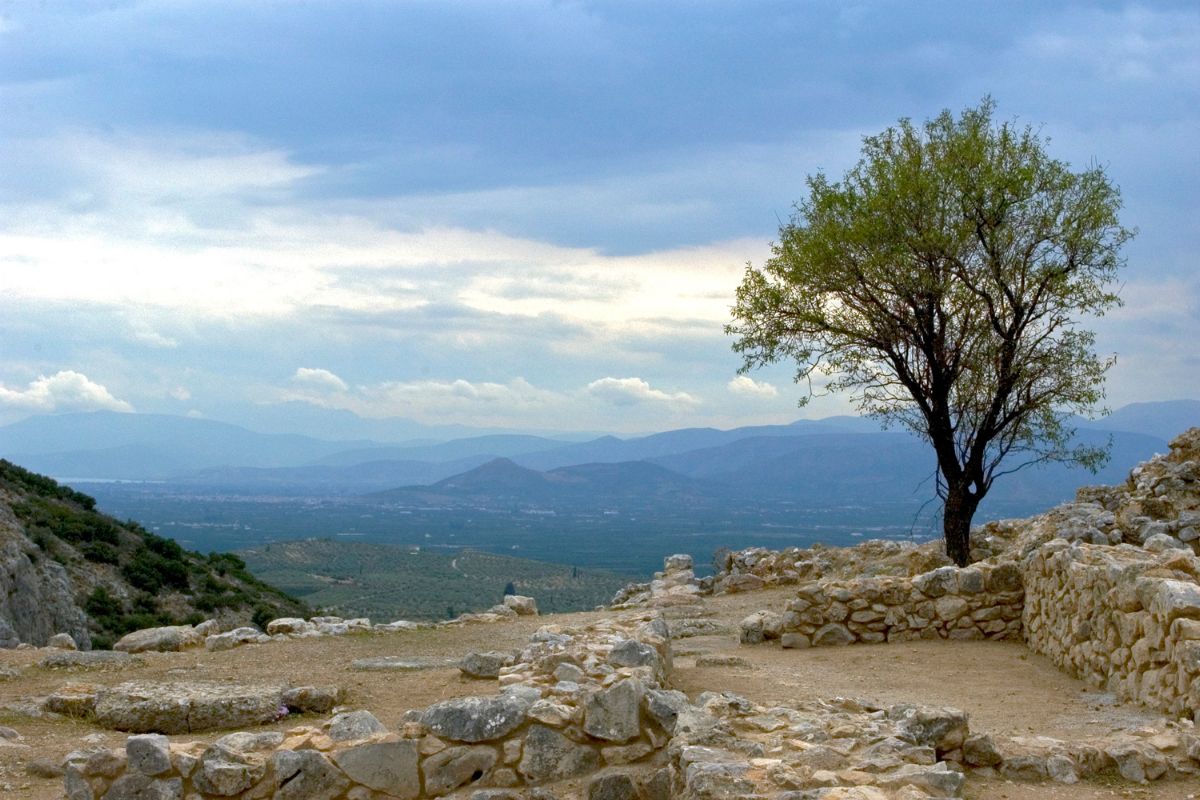 The image shows a view from the ancient site of Mycenae, featuring stone ruins in the foreground and a solitary tree, with a backdrop of rolling hills and a cloudy sky.