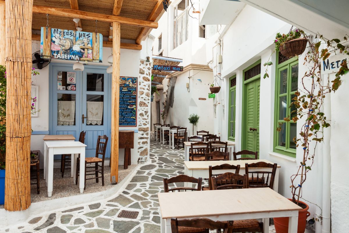 A cozy taverna in Naxos, with wooden tables and chairs set along a narrow, stone-paved alley, surrounded by whitewashed buildings adorned with colorful signs and potted plants.