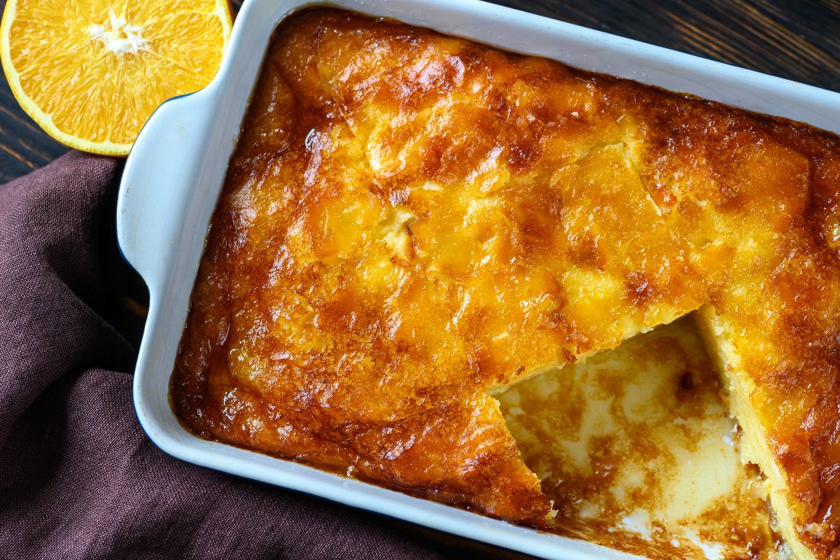 The image shows a tray of portokalopita, a traditional Greek orange phyllo pie. The dessert has a golden, caramelized top with a slice cut out, revealing its moist, syrupy interior. A halved orange is placed nearby for decoration.